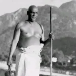 Swami Sivananda standing holding a wooden staff.
