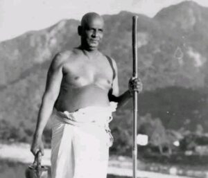 Swami Sivananda standing holding a wooden staff.