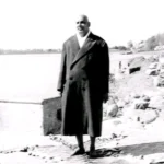 Swami Sivananda standing outdoors in long coat.