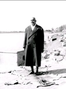 Swami Sivananda standing outdoors in long coat.