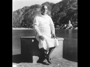 Sri Swami Sivananda seated peacefully beside a lake with mountains in the background