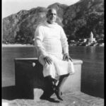 Sri Swami Sivananda seated peacefully beside a scenic lakeside with mountains