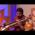 A musician in traditional red attire passionately playing the Saraswati Veena on stage