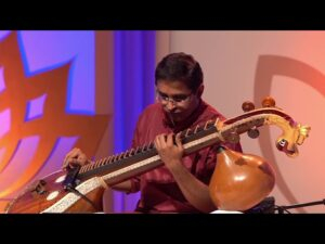 A musician in traditional red attire passionately playing the Saraswati Veena on stage